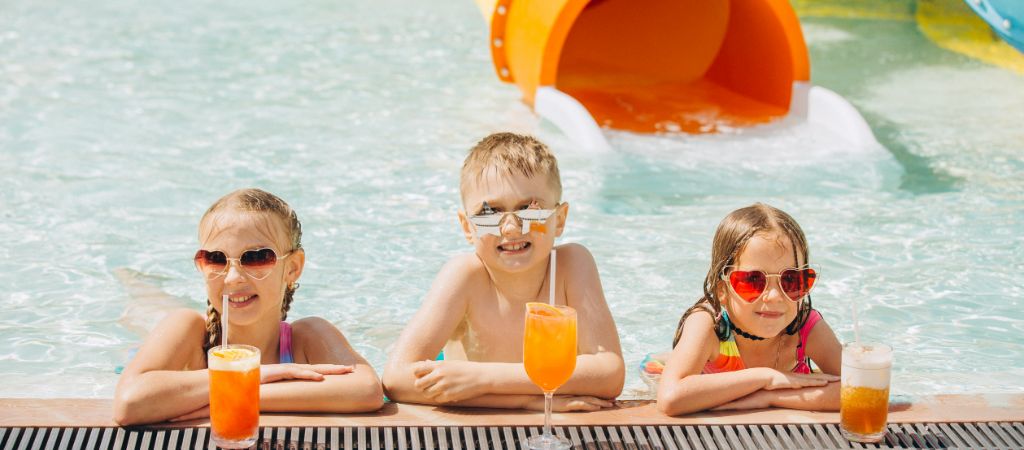 Children Enjoying Drinks at a Water Park Three kids in a pool by drinks, smiling near a water slide.